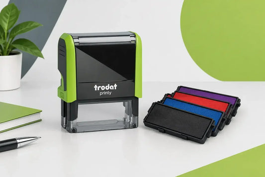 Trodot Printy self-inking stamp shown on a white desk with a plant, notebook, pen, and stacked ink pads in black, blue, purple, and red.