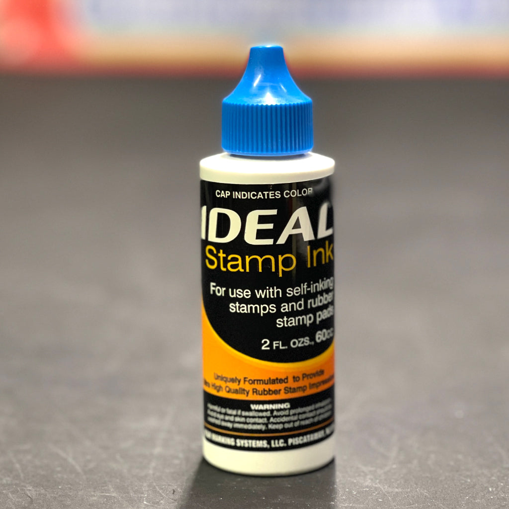 Bottle of IDEAL Stamp Ink with blue cap on a gray background