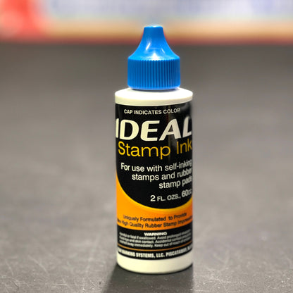 Bottle of IDEAL Stamp Ink with blue cap on a gray background