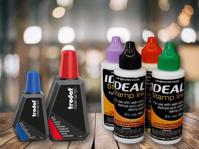 Collection of Ideal and Trodat stamp ink bottles on a wooden surface with a blurred background.
