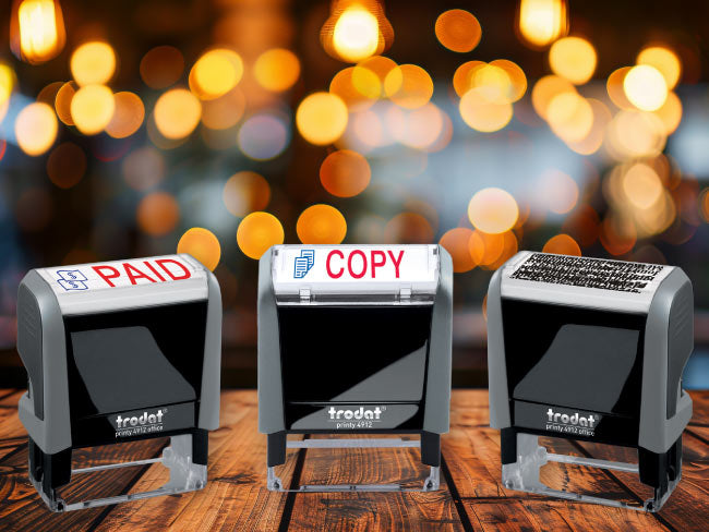 Three Trodat printy stamps labeled 'PAID', 'COPY', and 'CONFIDENTIAL' on a wooden surface with a blurred bokeh background.