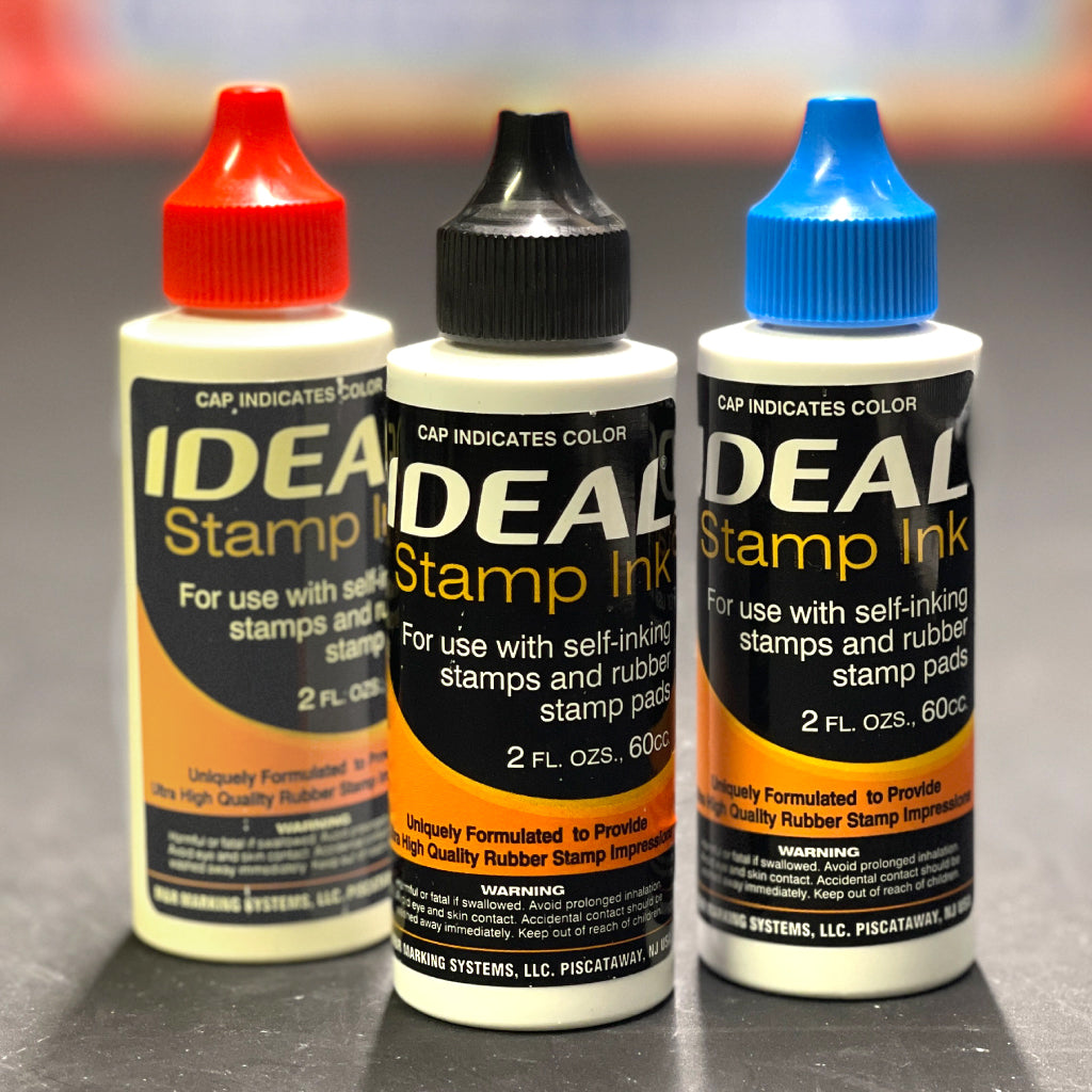 Three bottles of IDEAL Stamp Ink with different colored caps on a gray background