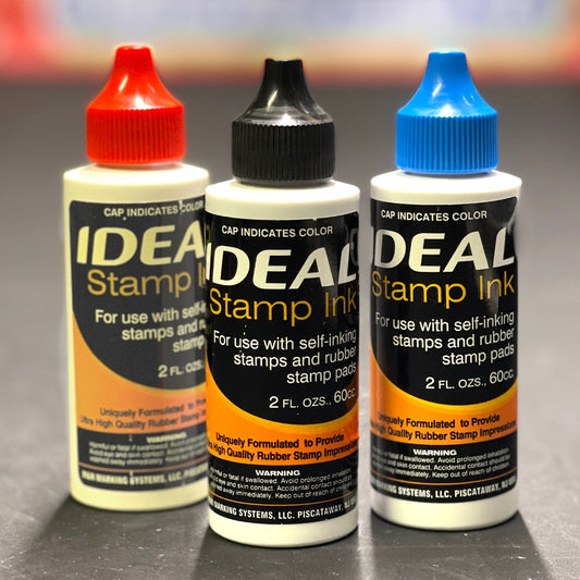 Three bottles of IDEAL Stamp Ink with different colored caps on a gray background