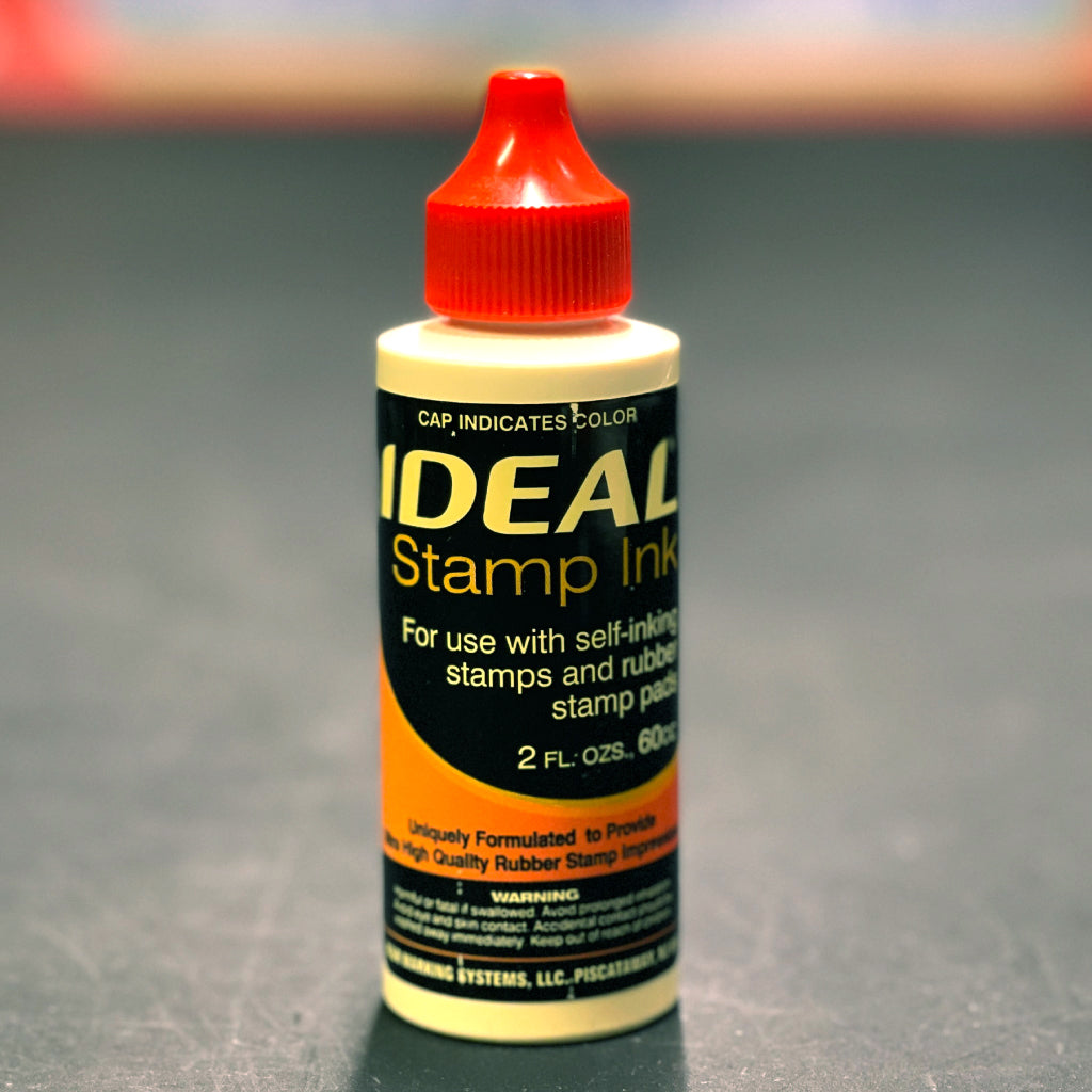 Bottle of IDEAL Stamp Ink with a red cap on a gray background